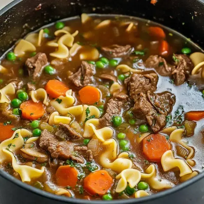 Une soupe épaisse contenant des pâtes, des morceaux de viande, des carottes et des petits pois dans un bouillon savoureux.