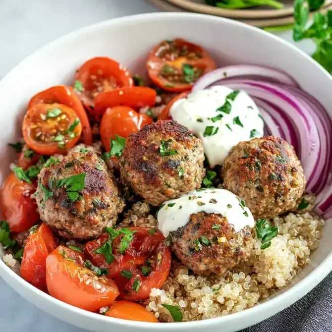 Un bol de quinoa garni de boulettes de viande, de tomates cerises, d'oignons rouges et de sauce blanche, le tout décoré de feuilles de coriandre.