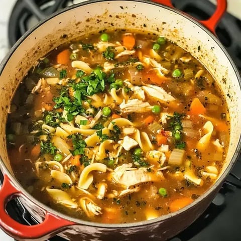 Une grande casserole de soupe de poulet aux nouilles, garnie de persil, avec des morceaux de poulet, des carottes, des petits pois et des oignons dans un bouillon savoureux.